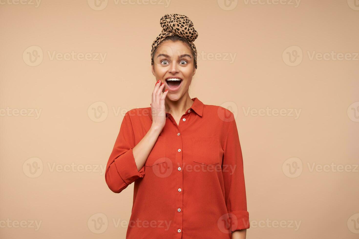 Bemused young lovely brunette female with natural makeup keeping palm on her cheek while looking surprisedly at camera with opened mouth, isolated over beige background photo