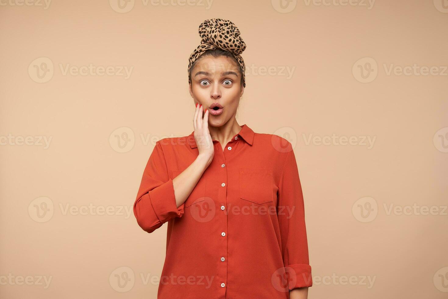 Dazed young attractive brown haired woman with headband raising surprisedly her eyebrows while looking amazedly at camera, standing over beige background photo