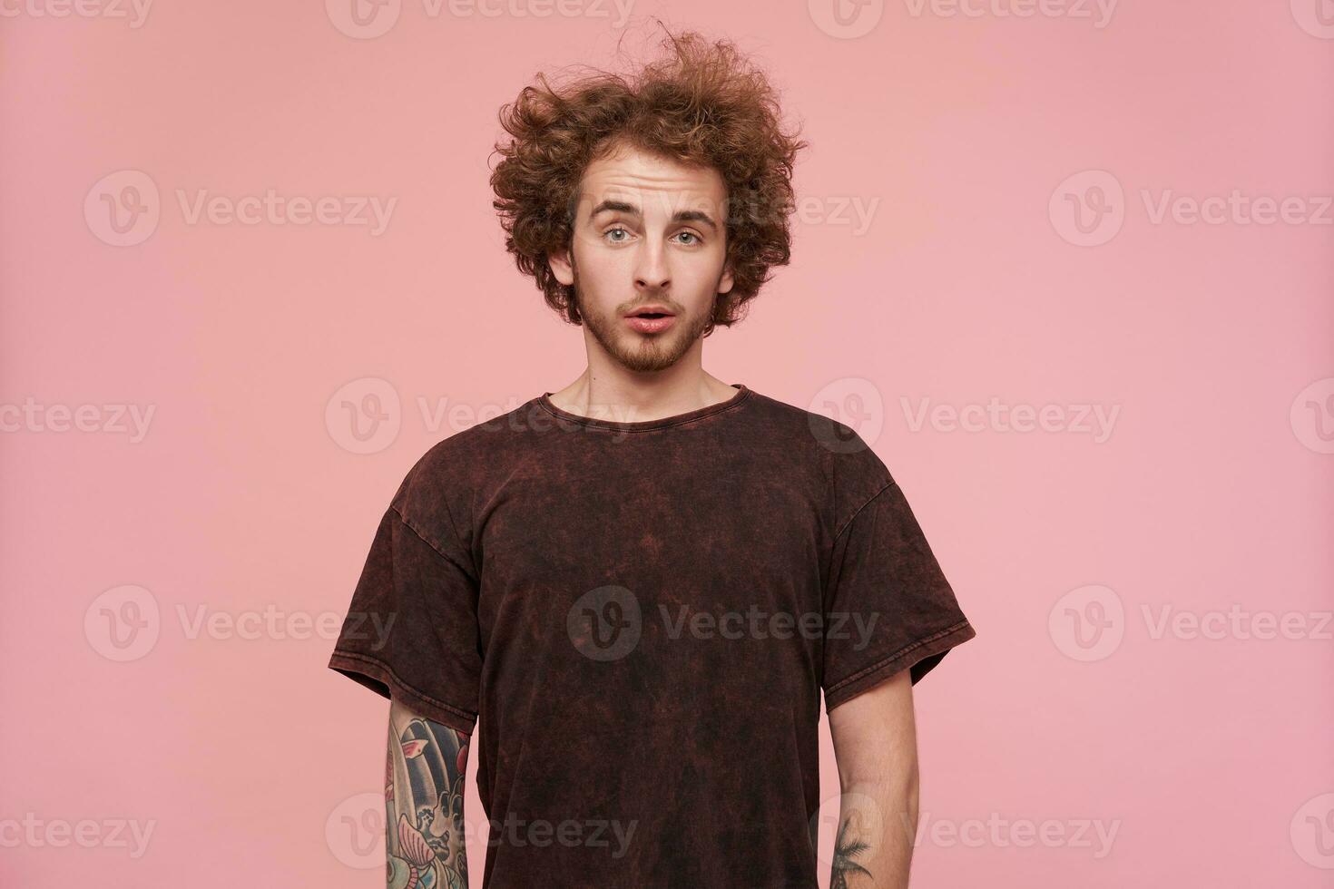Puzlled young handsome bearded redhead man with casual hairstyle looking confusedly at camera and wrinkling his forehead, standing against pink background photo