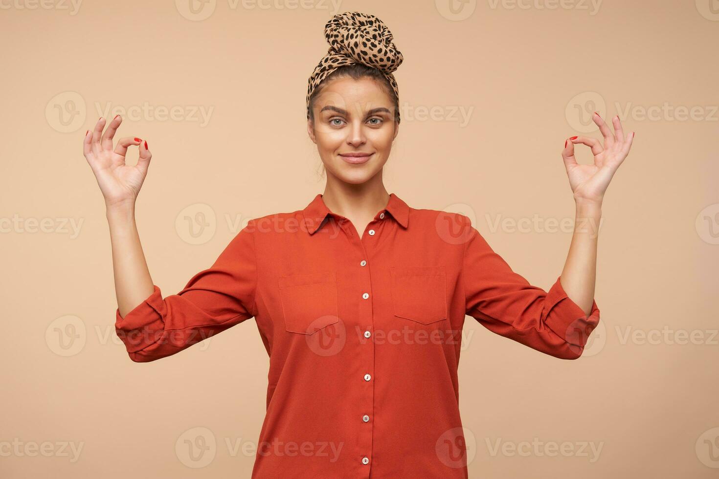 Positive young pretty brown haired woman with headband smiling slightly while looking at camera and folding fingers in mudra gesture, isolated over beige background photo