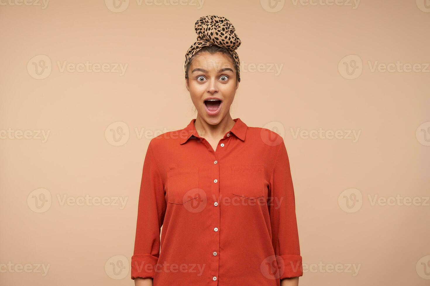 Open-eyed young attractive brown haired woman looking dazedly at camera with wide mouth opened, keeping hands down while standing over beige background photo