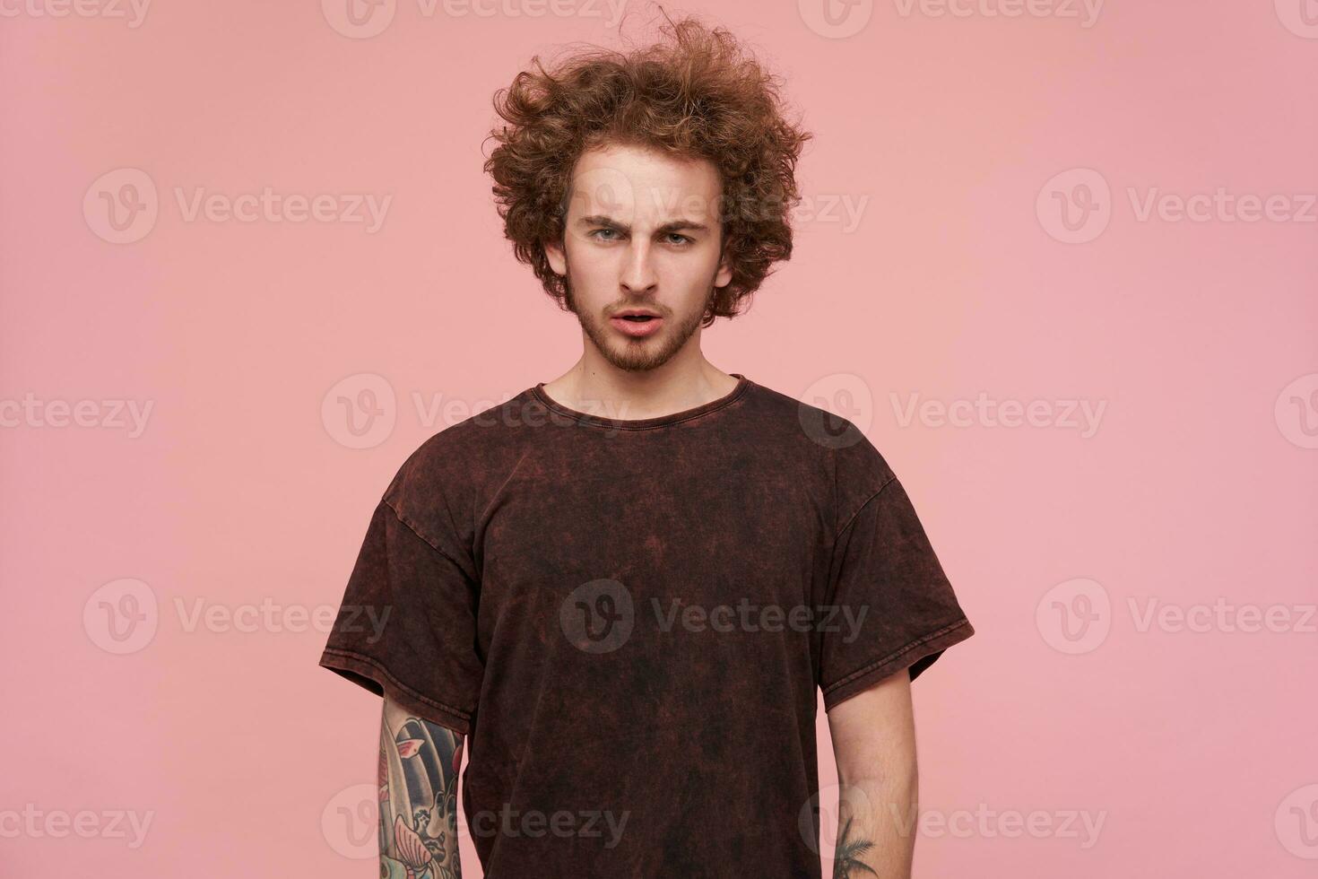 Confused young green-eyed curly bearded man frowning his eyebrows while looking seriously at camera, wearing dark red t-shirt while posing over pink background photo