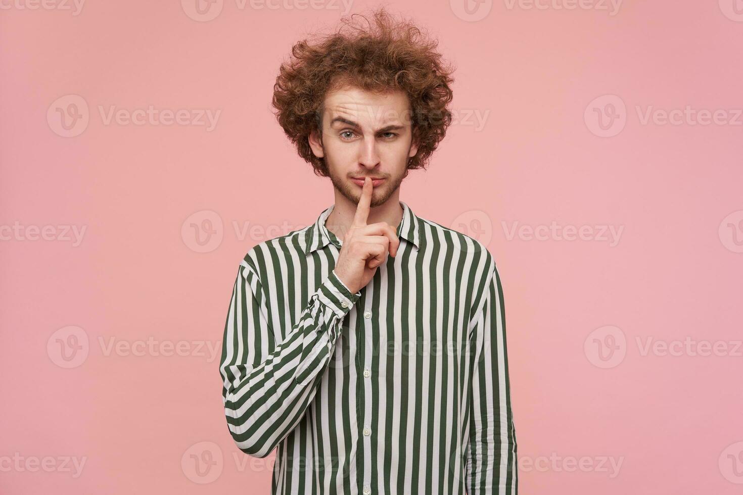 Portrait of lovely young curly redhead male dressed in casual clothes keeping forefinger on his mouth while looking at camera with raised eyebrow, standing against pink background photo