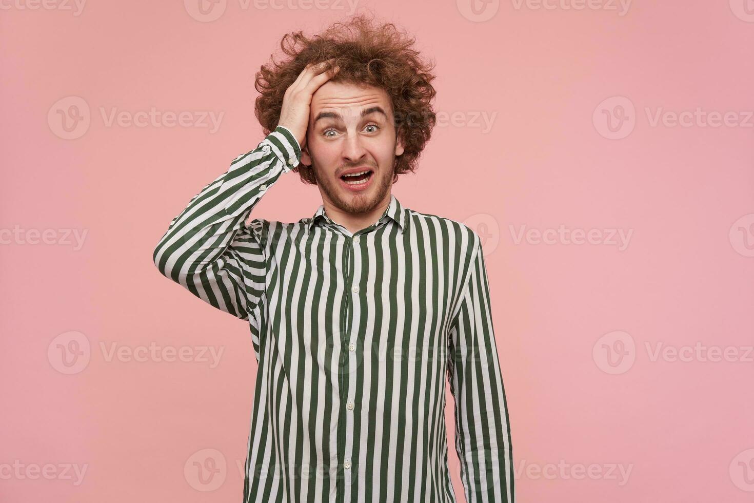 Open-eyed young pretty bearded redhead man with casual hairstyle wrinkling his forehead and keeping mouth opened while looking surprisedly at camera, posing over pink background photo