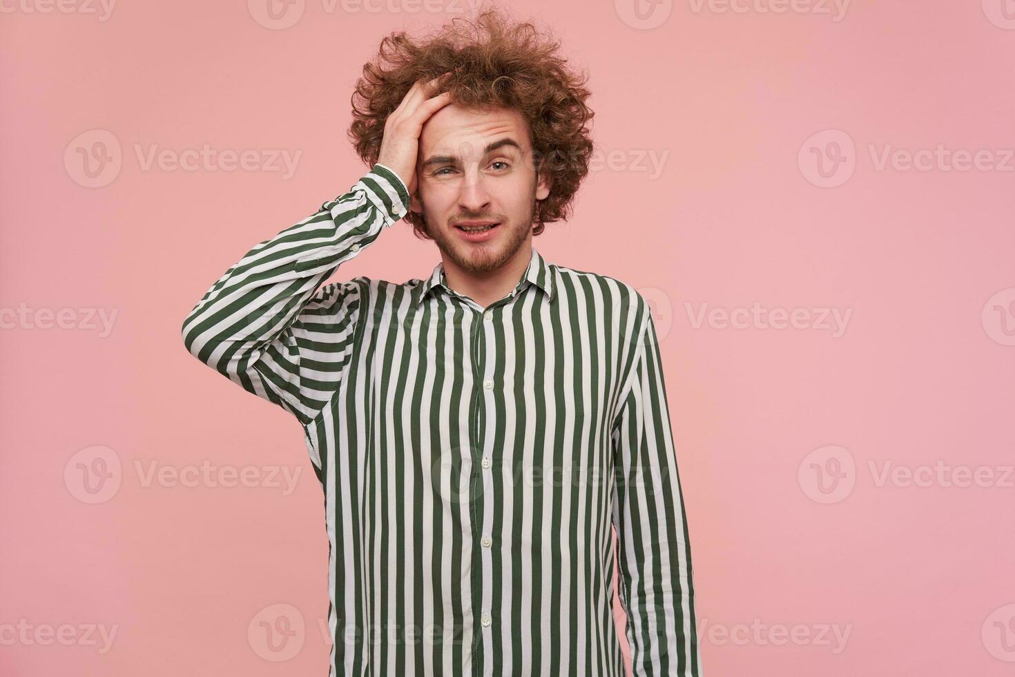 Indoor shot of confused young curly bearded male in striped shirt clutching his head while looking at camera, standing against pink background with puzzled face photo