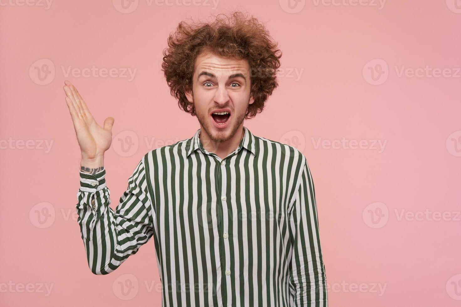 Angry young bearded curly male raising emotionally his palm while screaming crossly and frowning his face, dressed in striped shirt while standing over pink background photo