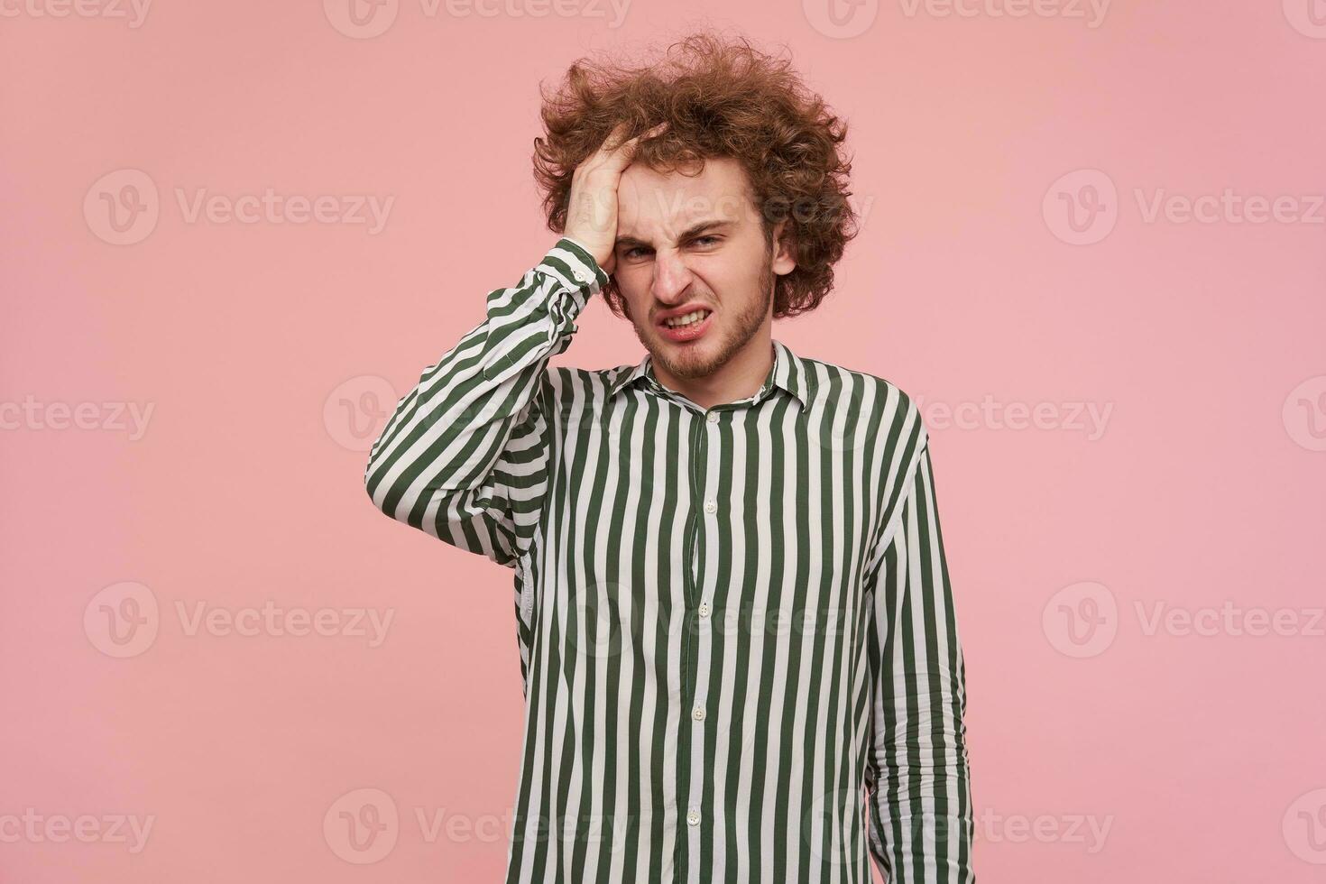 Indoor shot of displeased young bearded male clutching his curly red hair with raised handand looking at camera with pout, wearing striped shirt over pink background photo