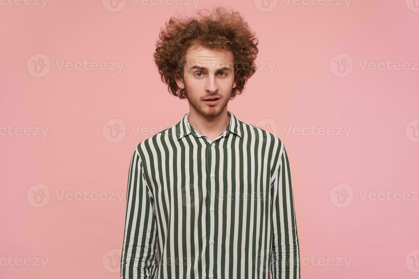 Attractive young curly redhead guy with casual hairstyle keeping hands down while posing over pink background, looking at camera with calm face and wrinkling forehead photo