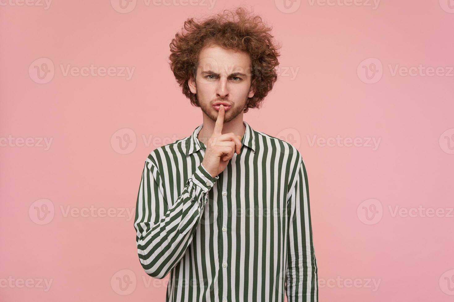 Studio photo of serious young bearded redhead male raising hand in hush gesture while posing over pink background, frowning his eyebrows while asking to keep silence