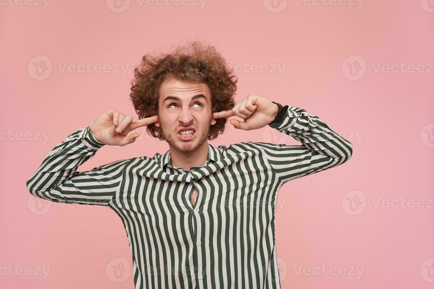 Displeased young bearded curly male with casual hairstyle inserting fingers into his ears while trying to avoid loud sounds, frowning face while posing over pink background photo