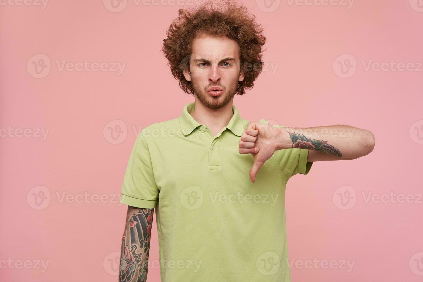 Displeased young curly brunette man with tattoos frowning eyebrows while looking discontentedly at camera while showing with thumb down, isolated over pink background photo