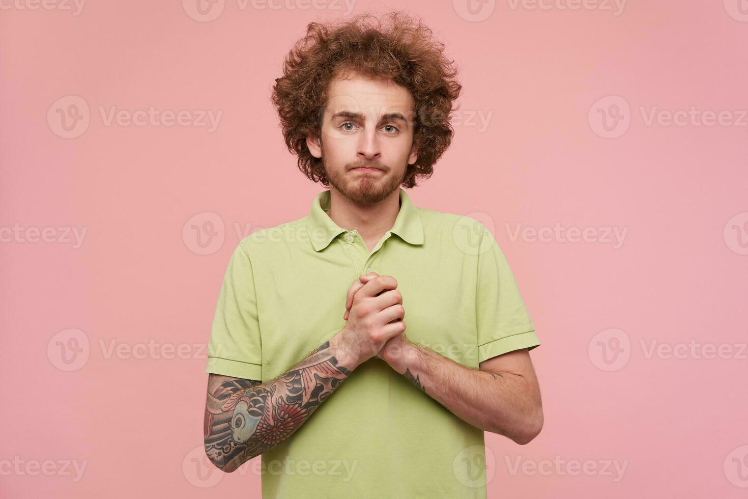 Indoor shot of young brunette guy with tattoos folding hands in praying gesture and looking desperately at camera, standing over pink background in casual wear photo