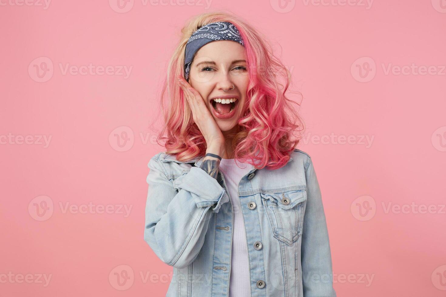 Portrait of happy cute girl with pink hair and tattooed hand,flattered by the compliment, laughs and touches the cheek standing over pink background, wearing a white t-shirt and denim jacket. photo