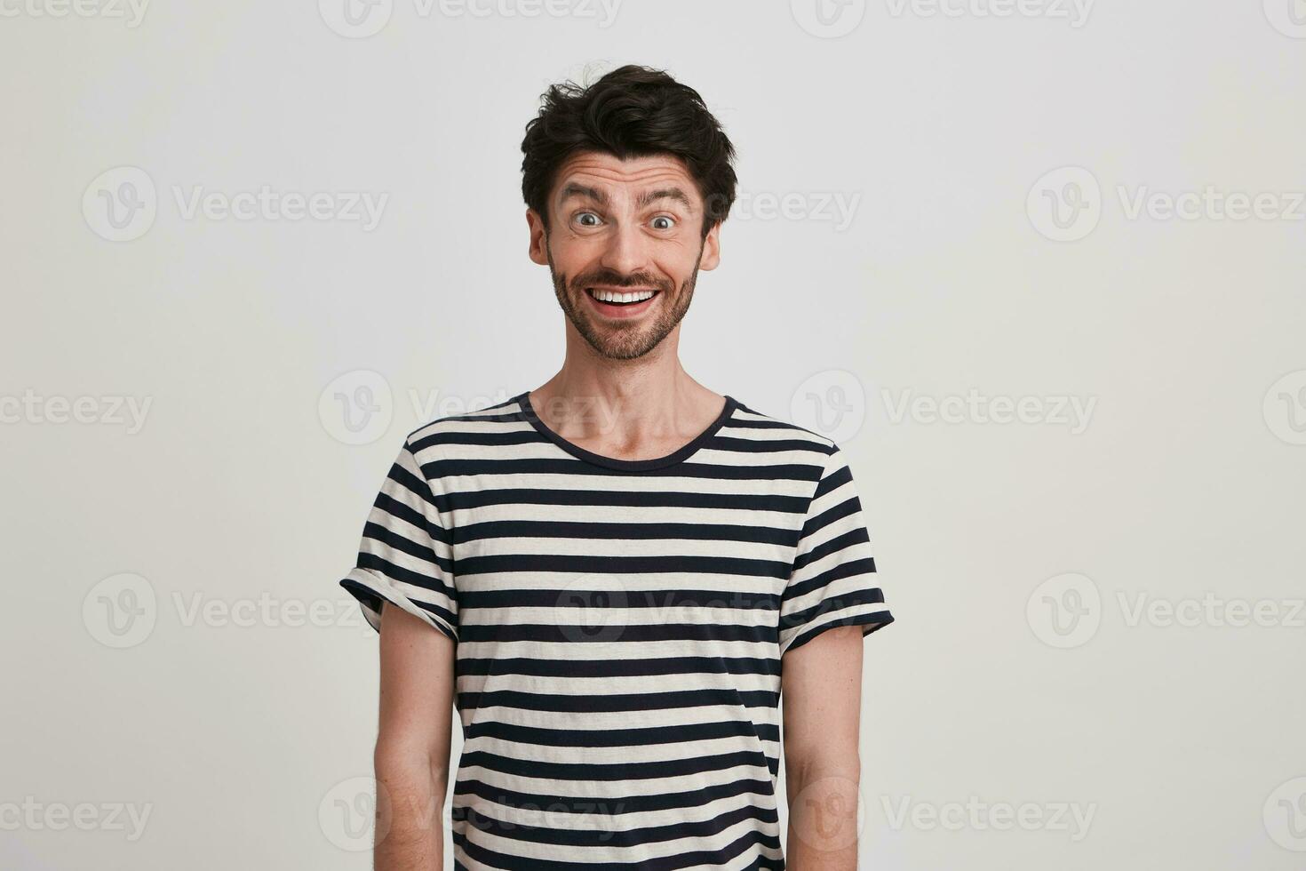 Portrait of cheerful amazed young man with bristle wears striped t shirt feels excited , standing and looks happy isolated over white background photo