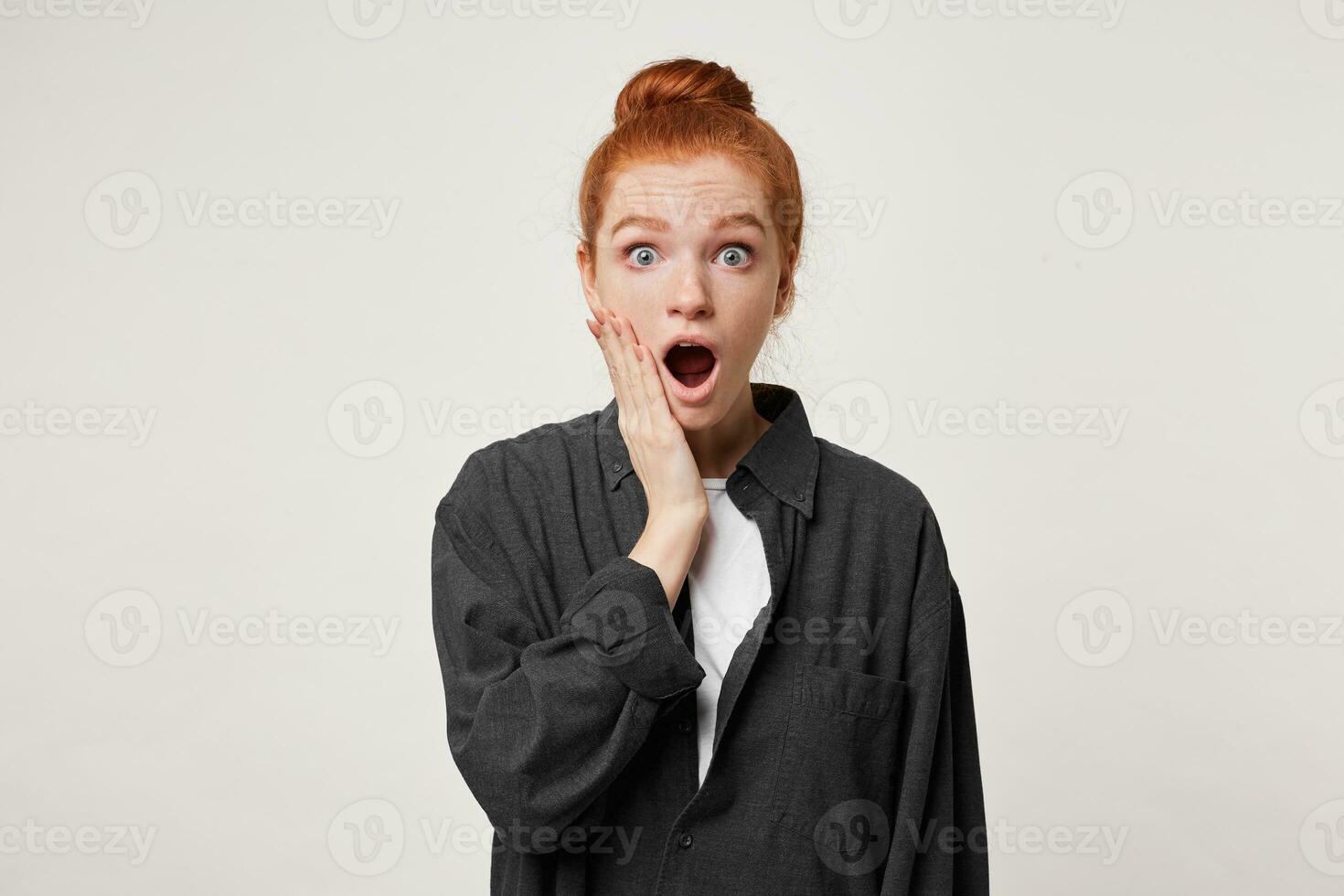 Red-haired girl stands in a daze, shocked by what she saw, with wide eyes looking at the camera, holding her hand near her cheek, her mouth open in surprise photo
