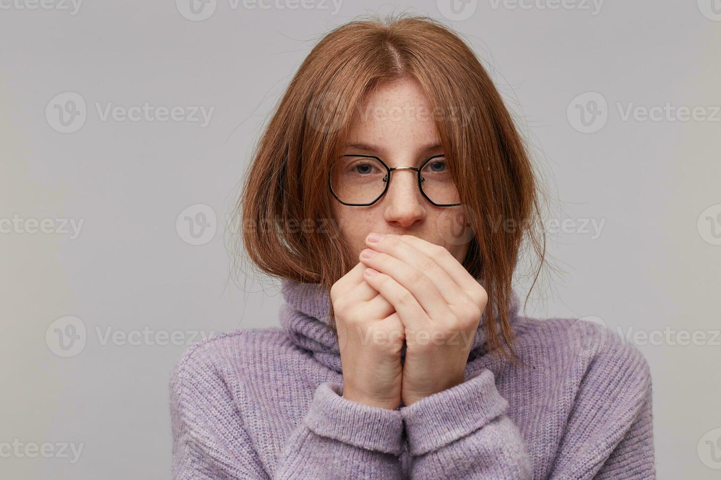 Young attractive red-haired girl in glasses dressed in a warm purple sweater with a high collar blowing on cold palms trying to get warm photo