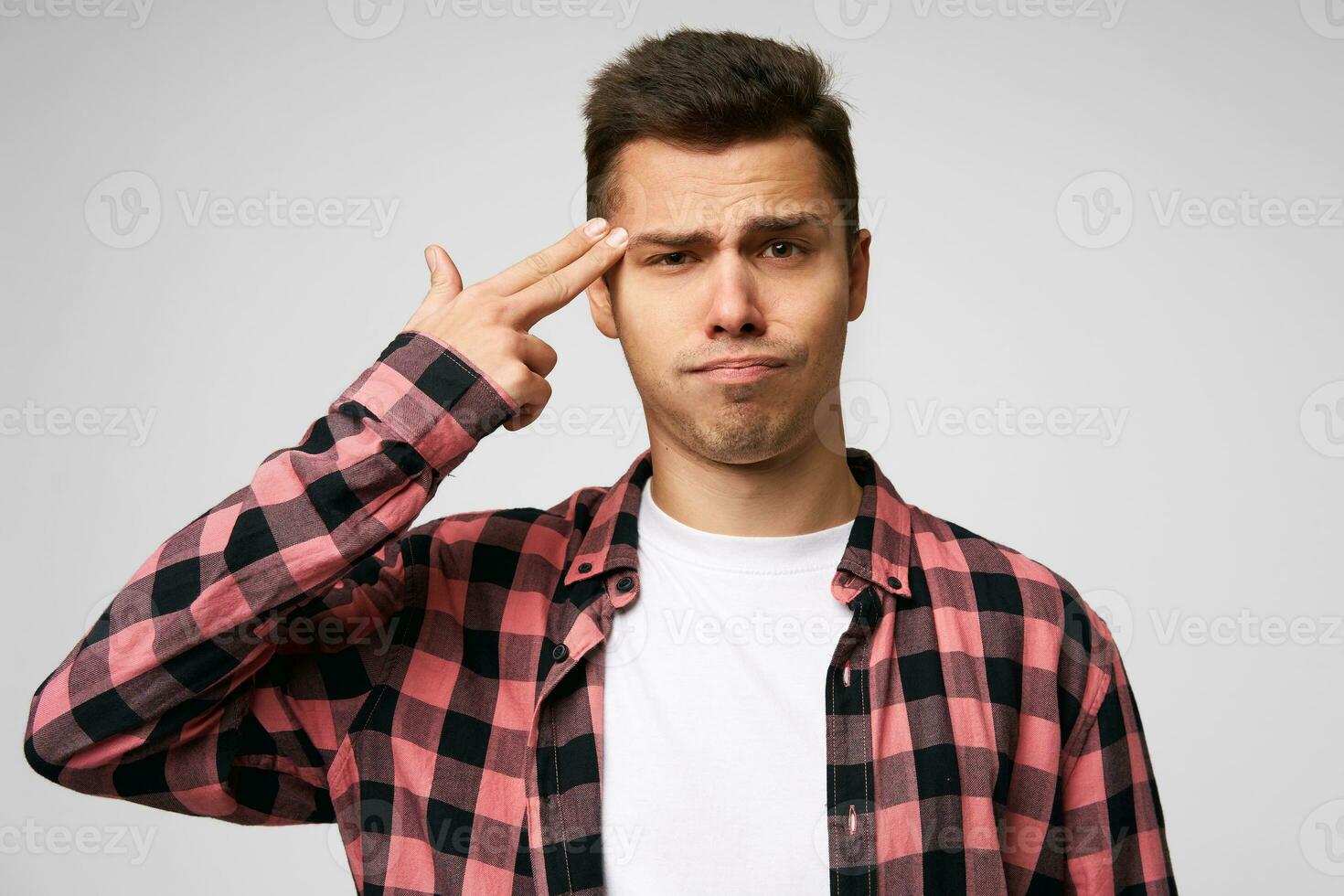 Unhappy man makes suicide gesture, being tired of everything, shoots in temple with hand, isolated over white studio background. Upset male gestures indoor, tries kill himself photo