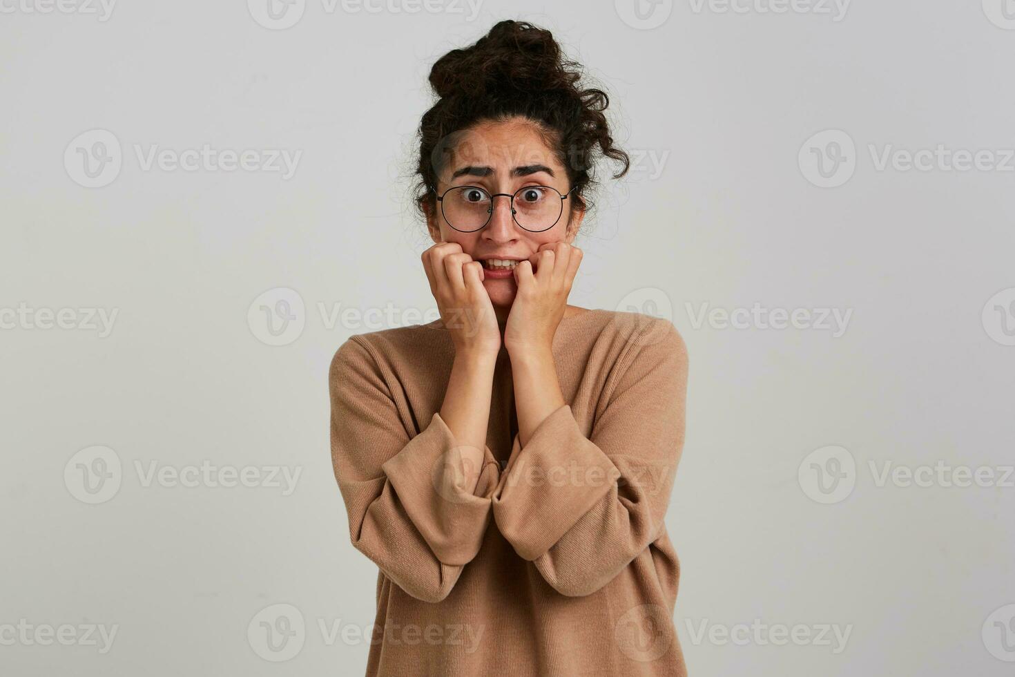 Portrait of horrified, adult girl with dark curly hair bun. Wearing beige jumper and glasses. Emotion concept. Touching her face in fear. Watching at the camera isolated over white background photo