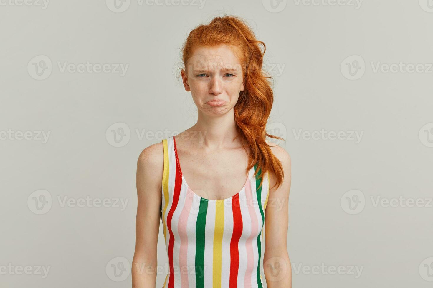 Portrait of unhappy, crying girl with ginger pony tail and freckles. Wearing striped colorful swimsuit. Emotion concept. Pouts her lip. Watching at the camera isolated over grey background photo