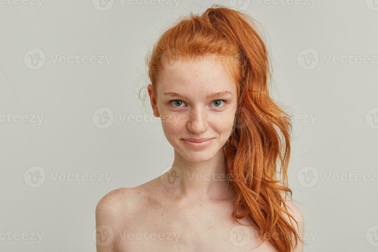 Nice looking woman, beautiful redhead girl with pony tail and freckles. Has no make up and stand shirtless. Emotion concept. Watching at the camera. Close up isolated over grey background photo