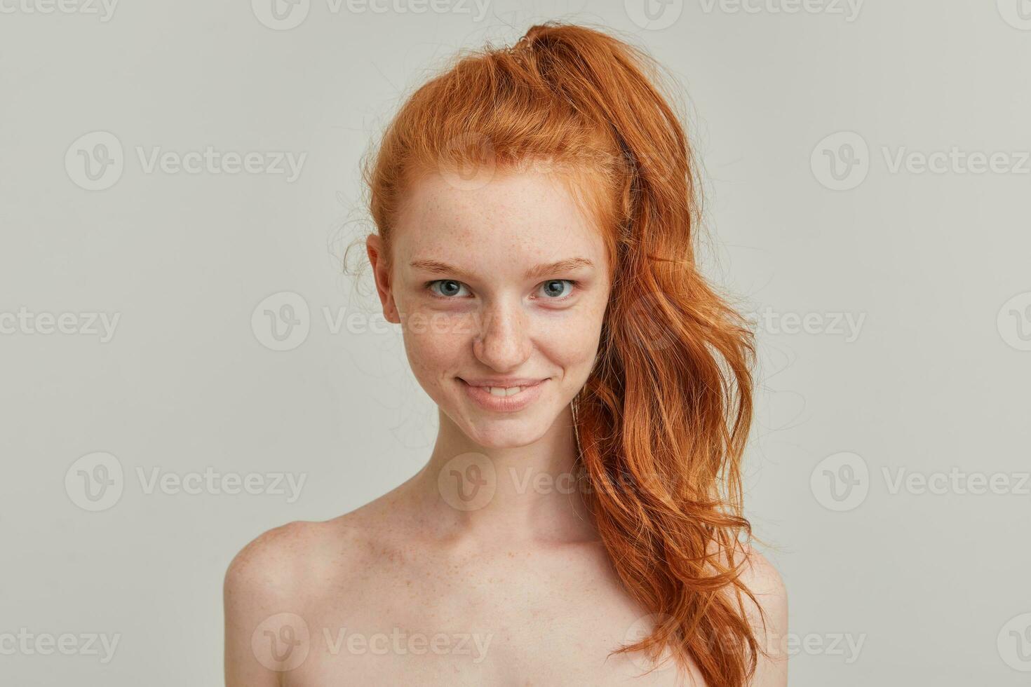 Young lady, pretty woman with ginger pony tail and freckles. Has no make up and stand shirtless. Emotion concept. Watching tempting at the camera, close up isolated over grey background photo