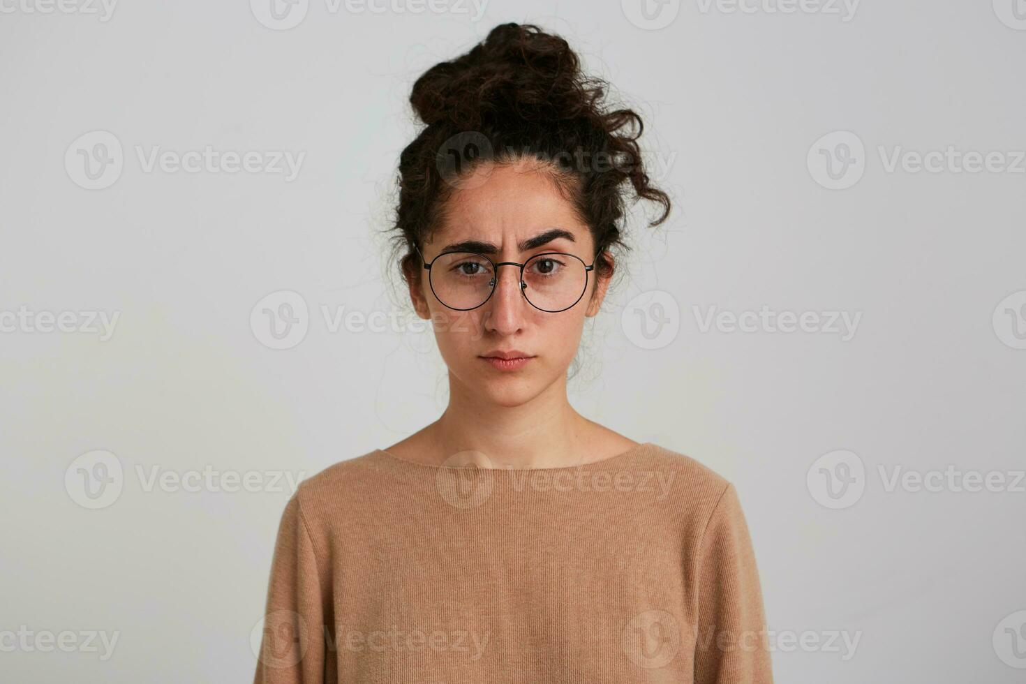 Frowning lady, serious woman with dark curly hair bun. Wearing beige jumper and glasses. Emotion concept. Lifts eyebrow. Watching unsure at the camera isolated over white background photo