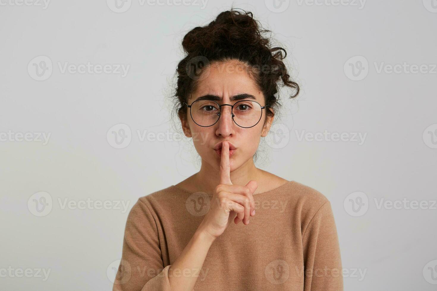 Portrait of unhappy, frowning girl with dark curly hair bun. Wearing beige jumper and glasses. Emotion concept. Showing silence sign, be quiet. Watching at the camera isolated over white background photo