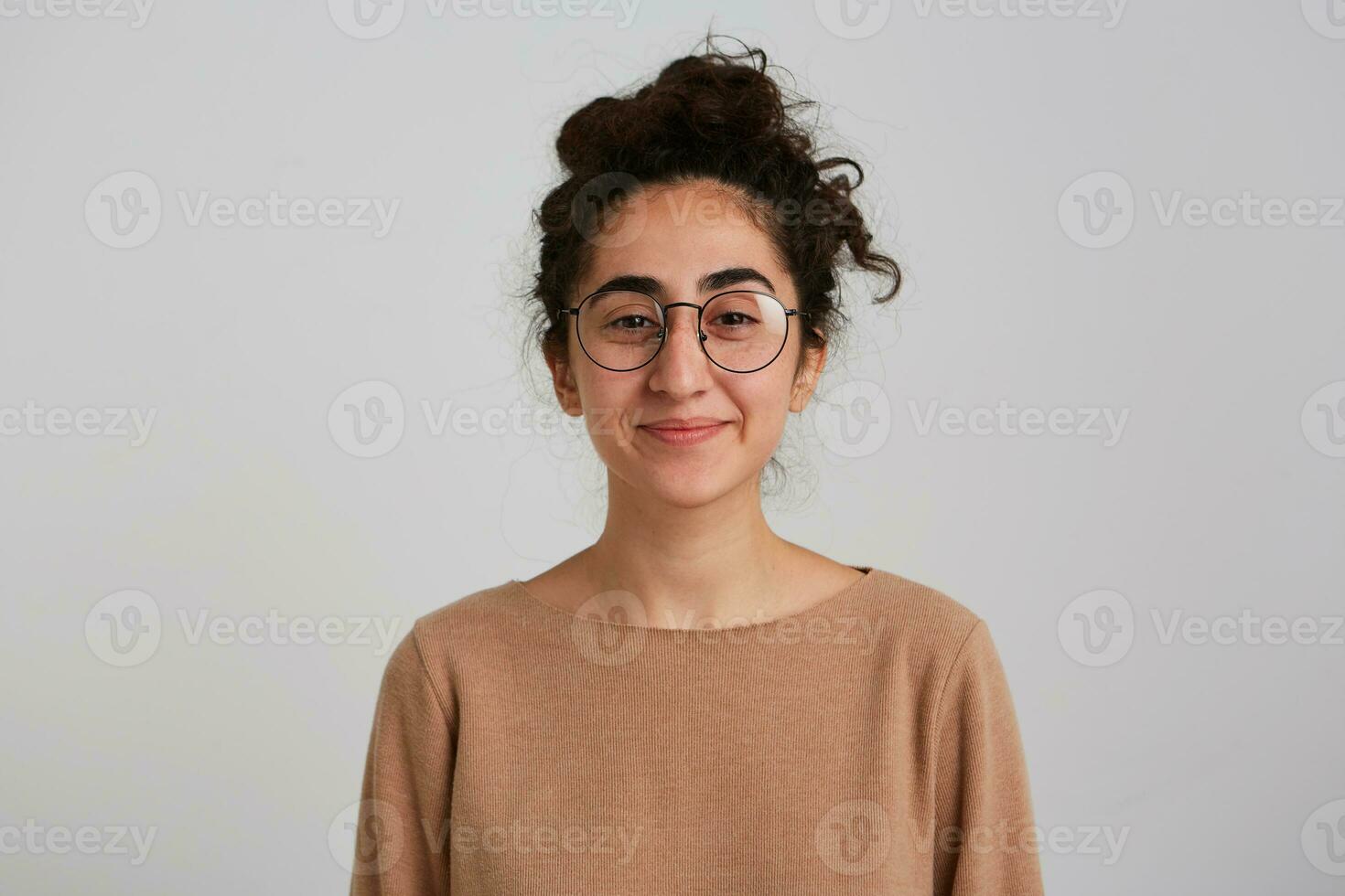 linda mirando mujer, hermosa niña con oscuro Rizado pelo bollo. vistiendo beige saltador y lentes. emoción concepto. acecho con sonrisa a el cámara aislado terminado blanco antecedentes foto