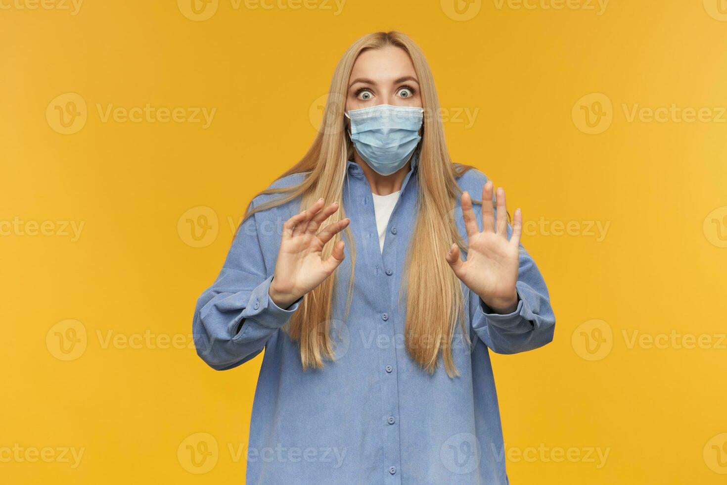 Scared looking woman, terrified girl with long blond hair. Wearing blue shirt and medical face mask. Pushes arms away, trying to protect. Watching at the camera, isolated over orange background photo