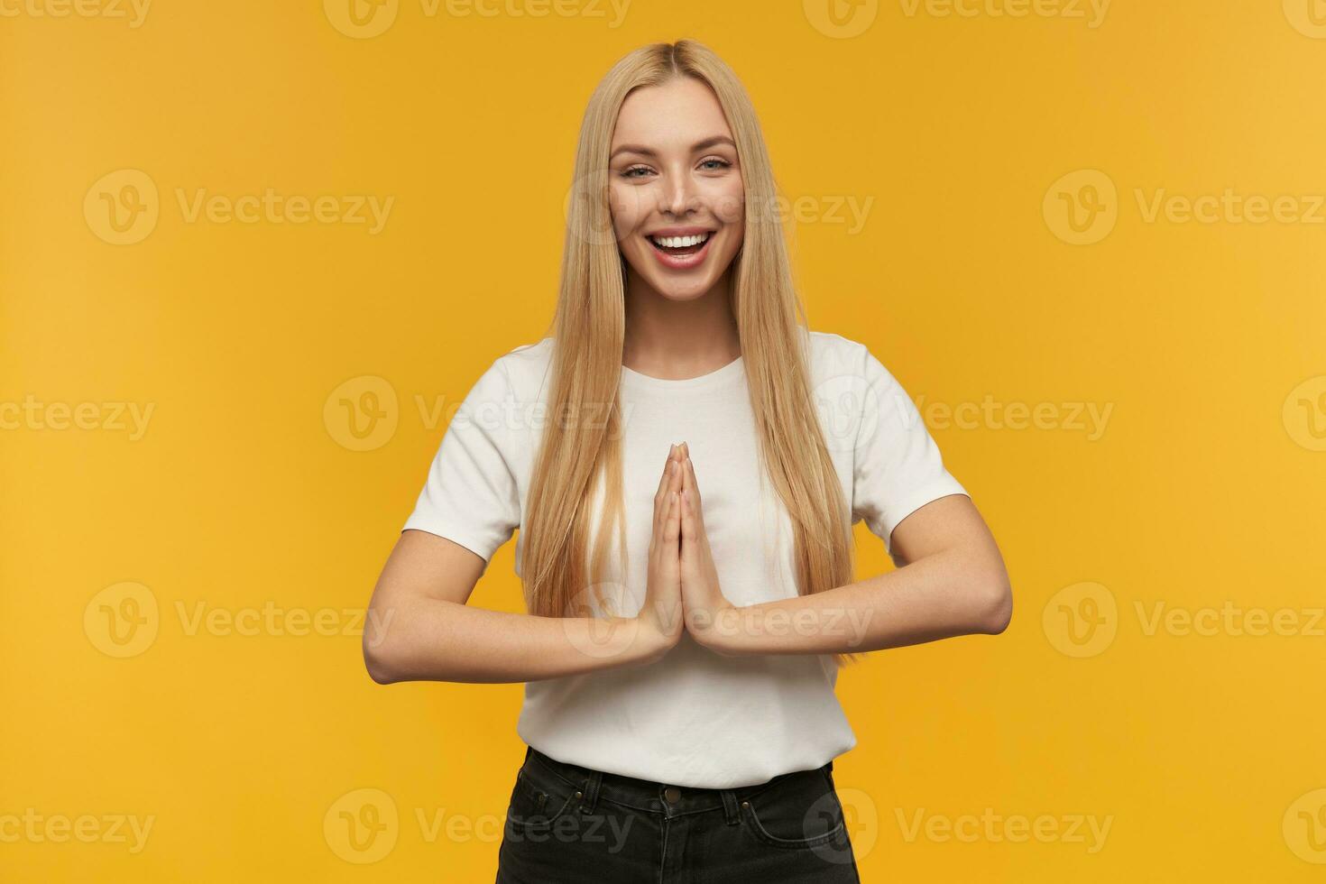 Portrait of attractive, adult girl with blond long hair. Wearing white t-shirt and black jeans. People and emotion concept. Stand isolated over orange background, keeps her palm together and praying photo