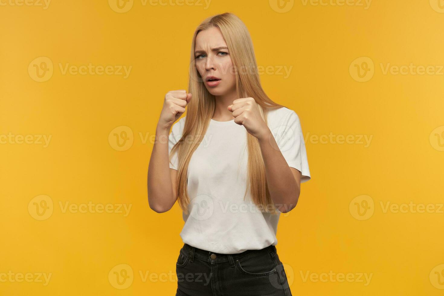 ready to fight, happy looking woman with blond long hair. Wearing white t-shirt and black jeans. People and emotion concept. Watching at the camera, isolated over orange background photo