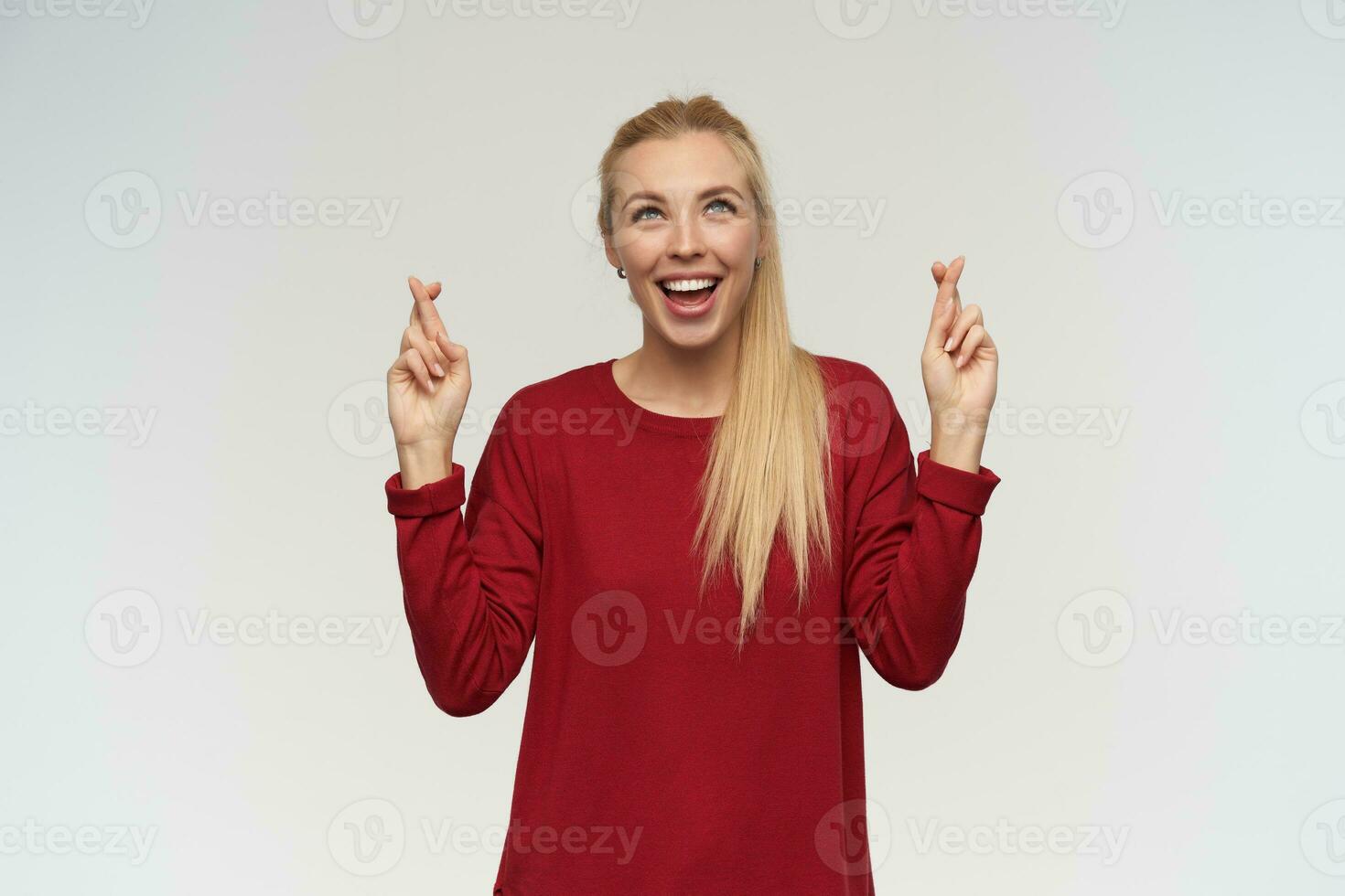 Portrait of attractive, adult girl with blond long hair. Wearing red sweater. People and emotion concept. Watching up at copy space, isolated over white wall, keeps her fingers crossed and praying photo