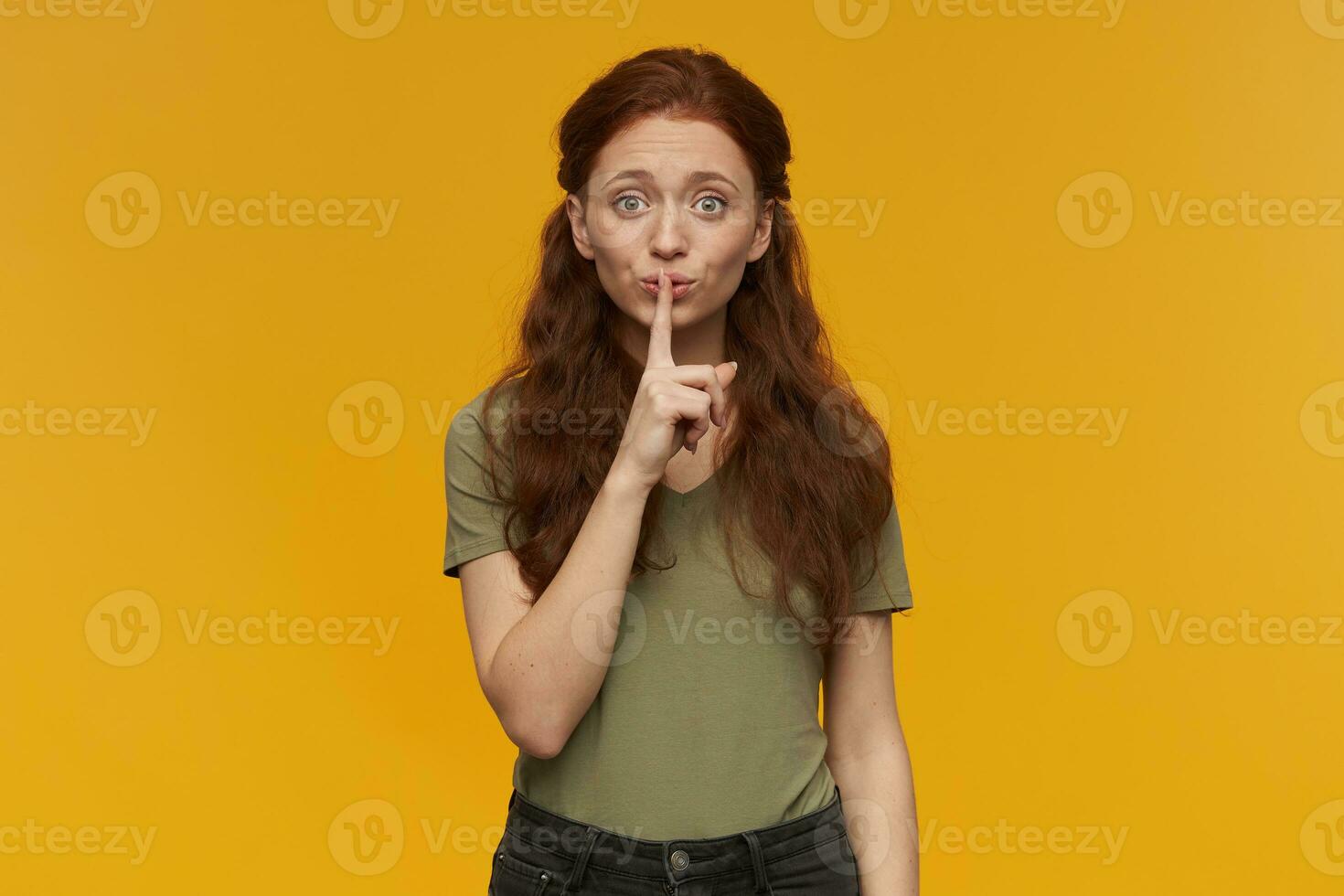 Happy looking girl, attractive redhead woman with long hair. Wearing green t-shirt. Emotion concept. Showing silence sign, ask to be quiet. Watching at the camera, isolated over orange background photo