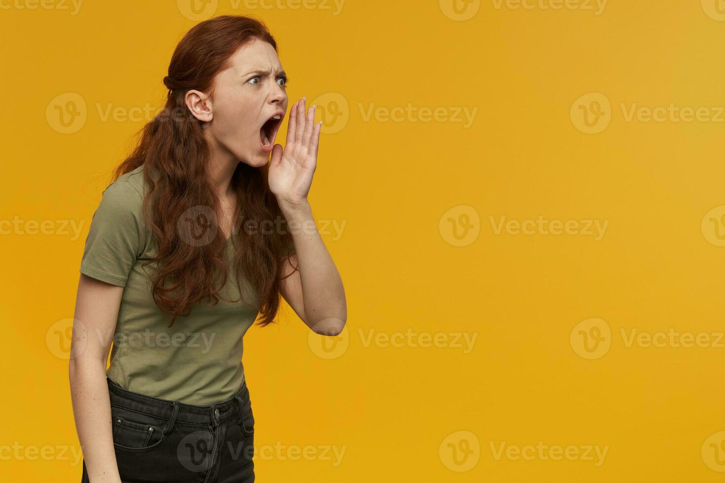Young, frowning woman with long ginger hair. Wearing green t-shirt. Emotion concept. Holding palm next to her mouth and shout. Watching to the right at copy space, isolated over orange background photo