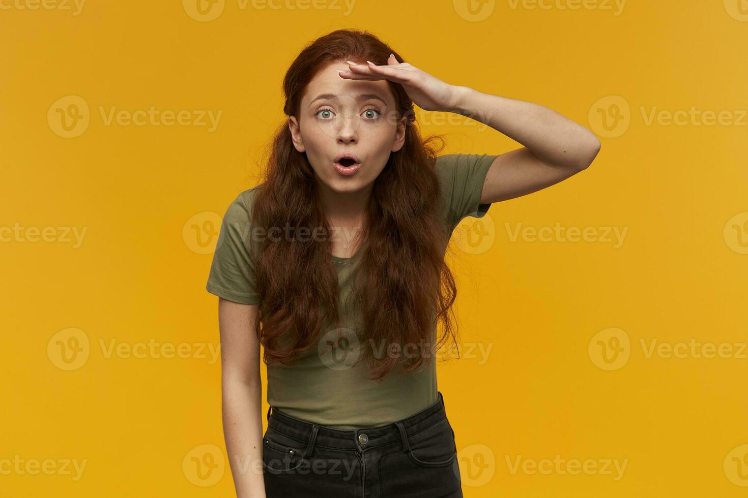 Surprised, positive woman with long ginger hair. Wearing green t-shirt. People and emotion concept. Gaze into distance with palm over her eyes. Watching at the camera, isolated over orange background photo