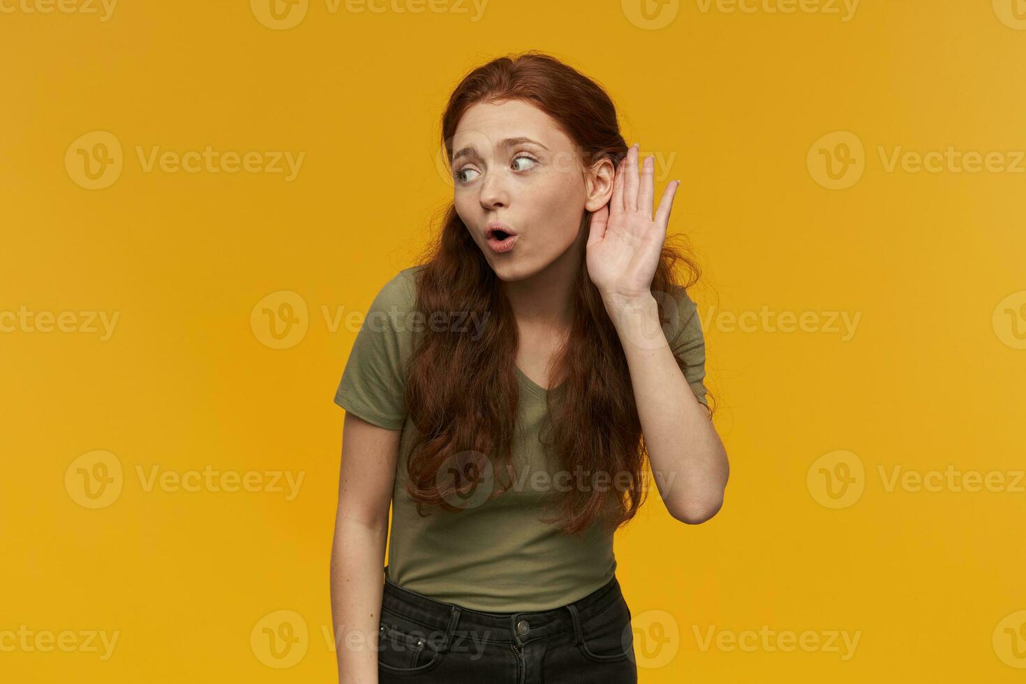 Portrait of curious, beautiful lady with long ginger hair. Wearing green t-shirt. People and emotion concept. Keeps palm next to ear, eavesdrop conversation. Stand isolated over orange background photo