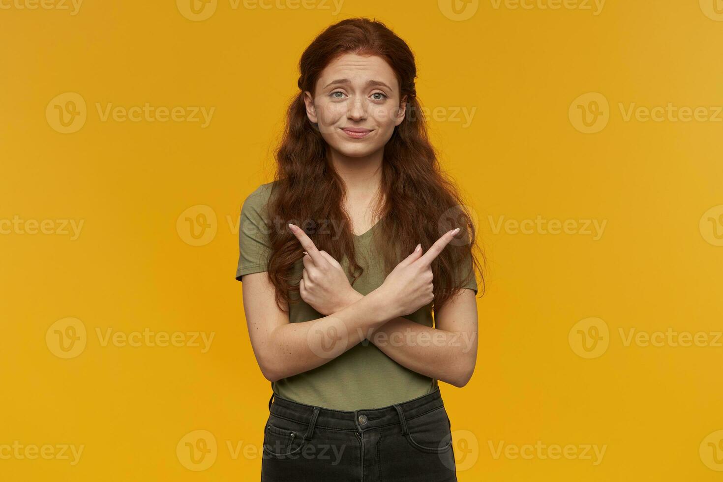 Cute, positive woman with long ginger hair. Wearing green t-shirt. People and emotion concept. Pointing uncertainly both sides at copy space. Watching at the camera, isolated over orange background photo