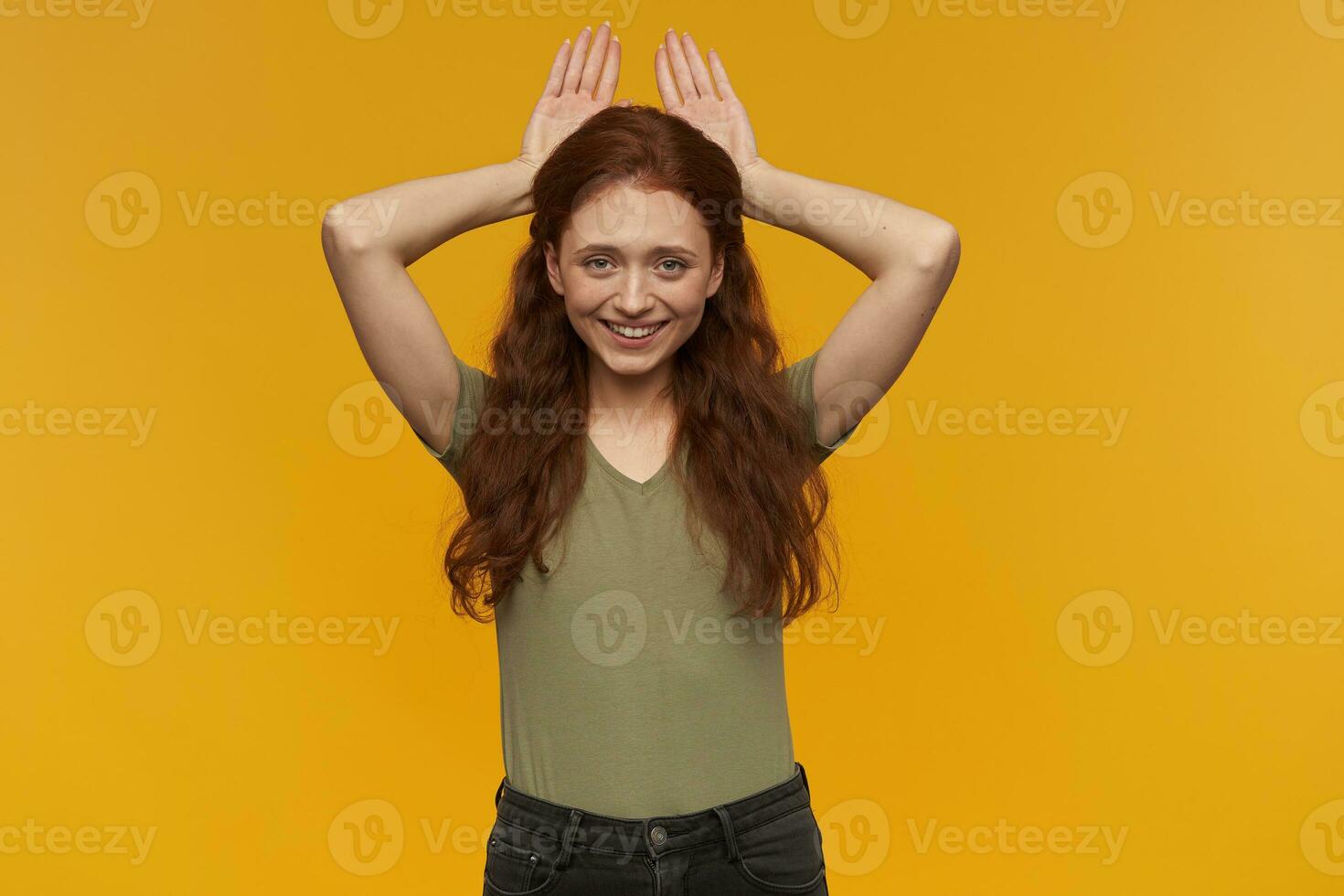 Portrait of playful, beautiful lady with long ginger hair. Wearing green t-shirt. Holding palms behind head like she has bunny ears. Watching at the camera, isolated over orange background photo