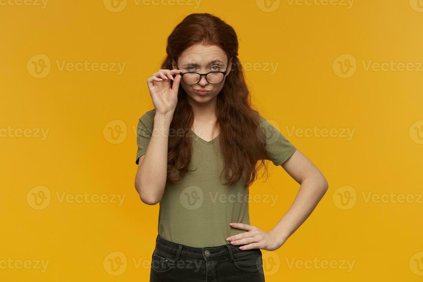 lindo, coqueta mujer con largo jengibre cabello. vistiendo verde camiseta y lentes. personas y emoción concepto. conmovedor su gafas y ascensores ceja. acecho a el cámara, aislado terminado naranja antecedentes foto