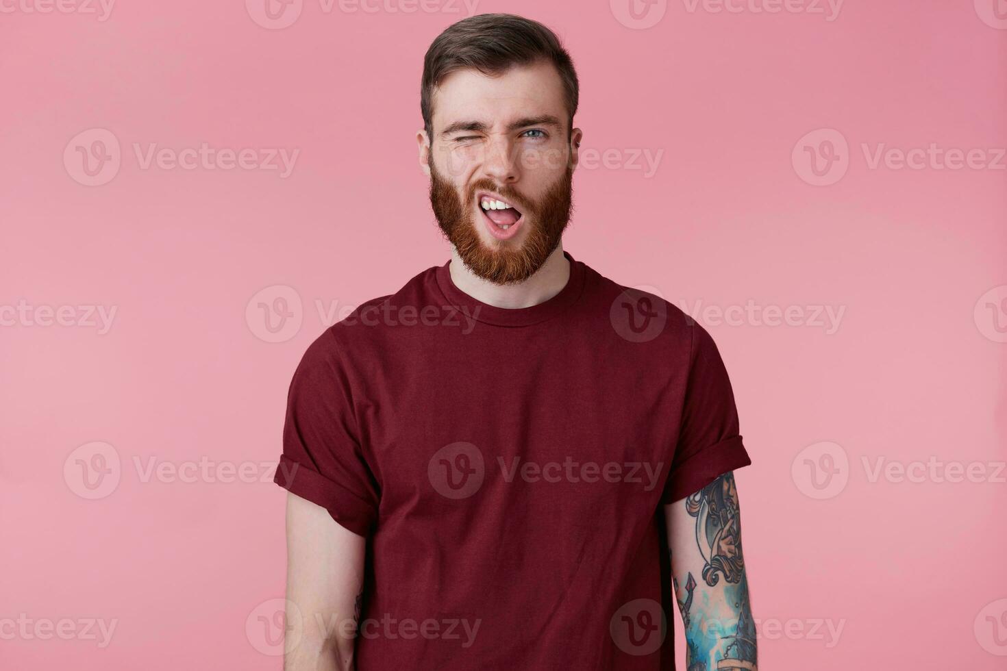 Portrait of young bearded male with tattooed hand makes a frown, screwing eye, isolated over pink background. photo