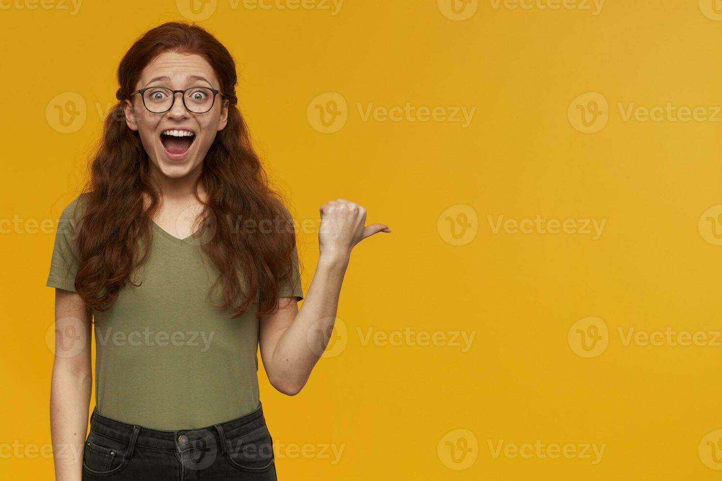 Excited, amazed woman with long ginger hair. Wearing green t-shirt and glasses. Watching at the camera and pointing with thumb to the right at copy space, isolated over orange background photo