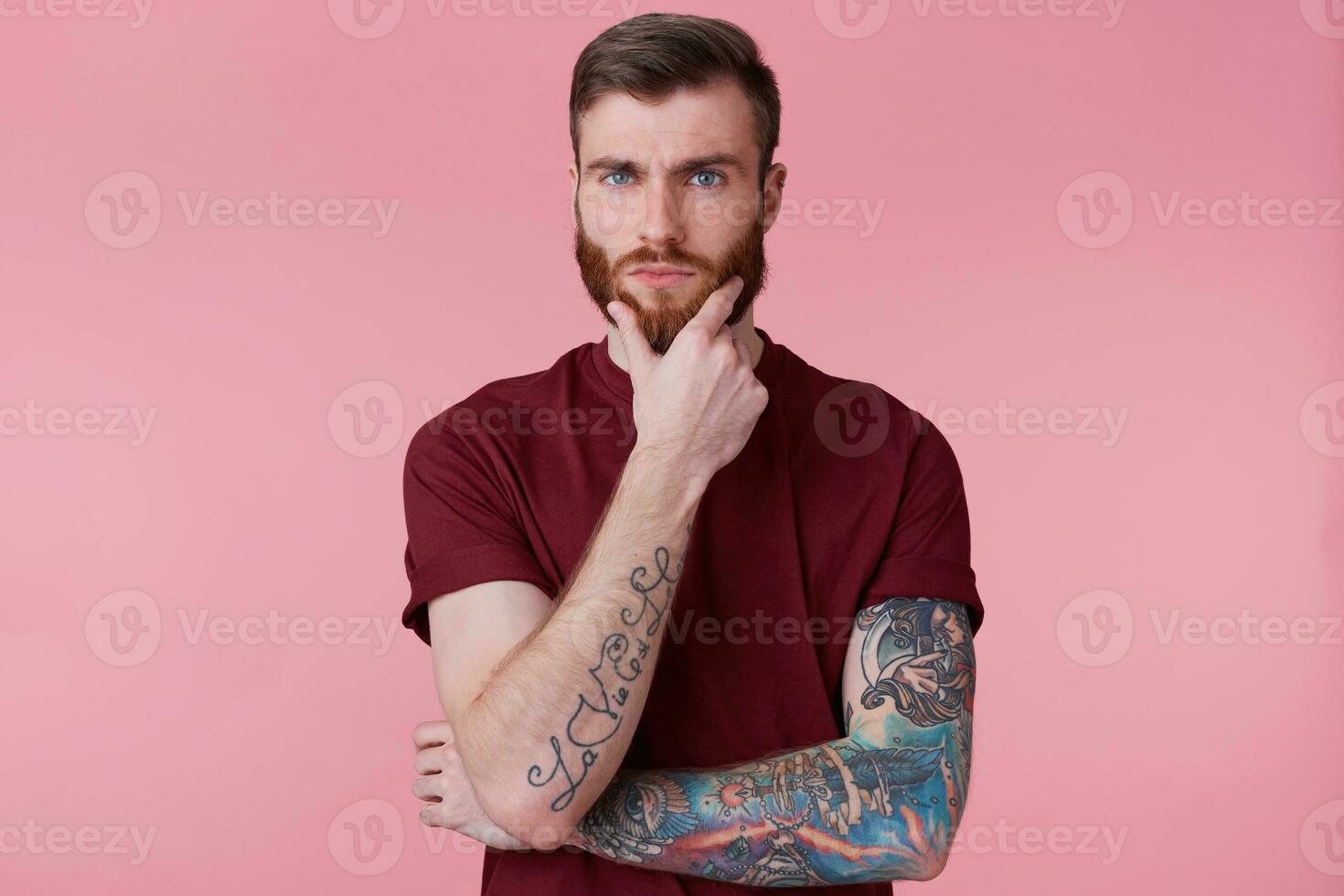 Close up of handsome thoughtful bearded guy with tattooed hand, looking at camera, holding his chin, thinks about his future, make plans, dreams, isolated over pink background. photo