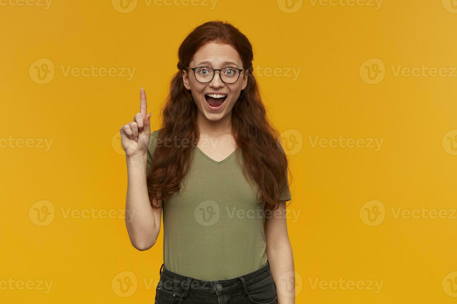 Cute, positive woman with long ginger hair. Wearing green t-shirt and glasses. People and emotion concept. Raises index finger up, got an idea. Watching at the camera, isolated over orange background photo