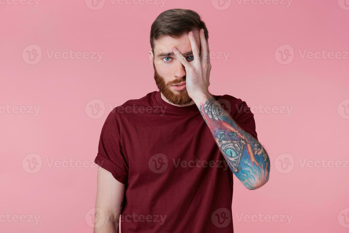 Photo of tired and disappointed young bearded man with tattooed hand, covers part of face with hand, looking through fingers isolated over pink background.