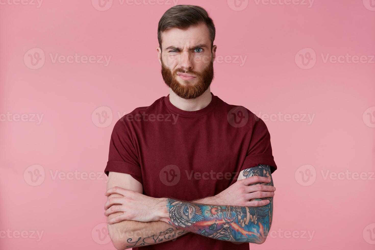 Photo of displeased beautiful bearded young man with tattooed hand, holding hands crossed, frowning, looking at camera isolated over pink background. People and emotion concept.