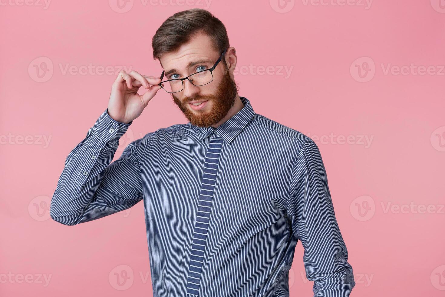 Close up of young handsome bearded man posing at camera isolated over pink background and smiling, holding his glasses with hand and looking through them. photo