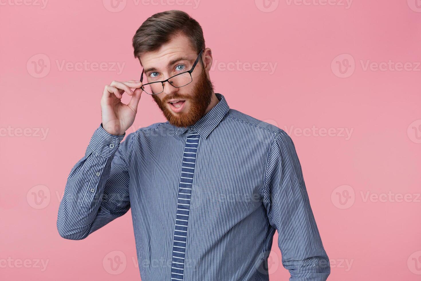 Close up of young successful man looks to the camera through his glasses isolated over pink background, open mouth, a little surprised. photo
