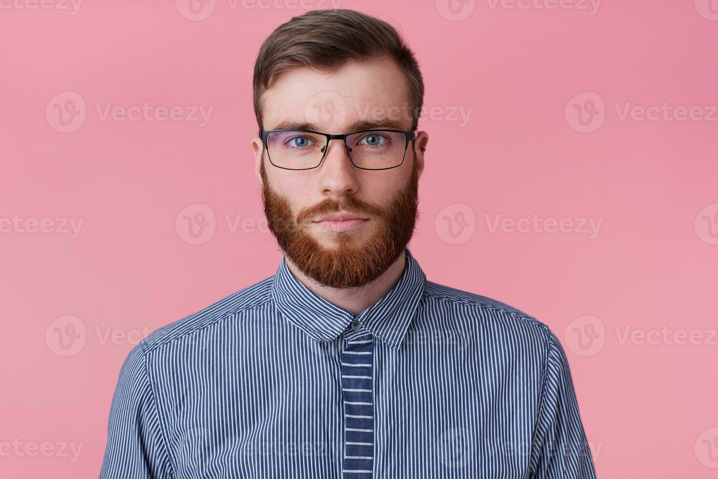 Close up of calm emotionless bearded young man in glasses, looking at the camera without emotion isolated over pink background. photo