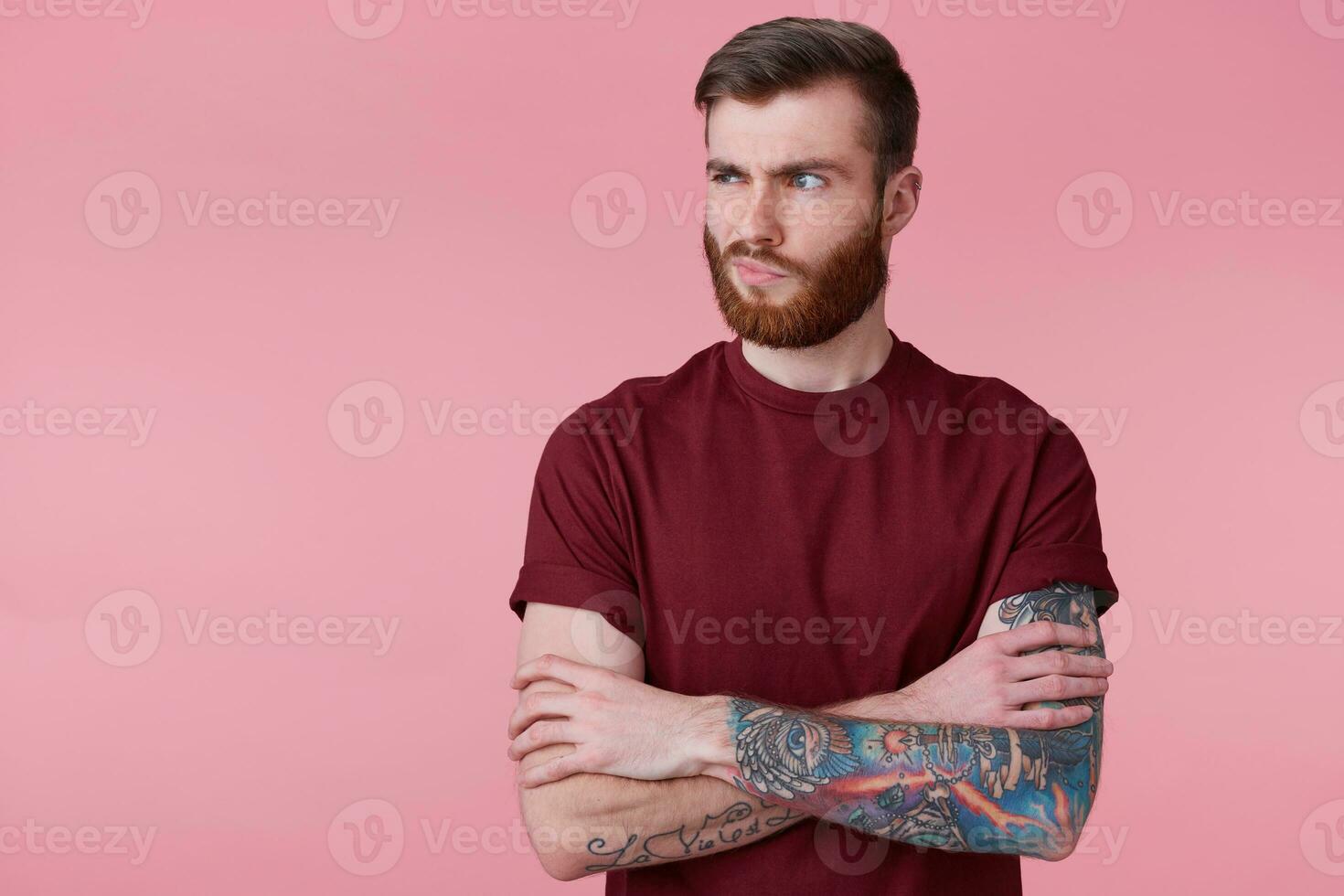 Portrait of displeased beautiful young man with ginger beard and tattooed hand, holding hands crossed, frown and looking away isolated over pink background. People and emotion concept. photo