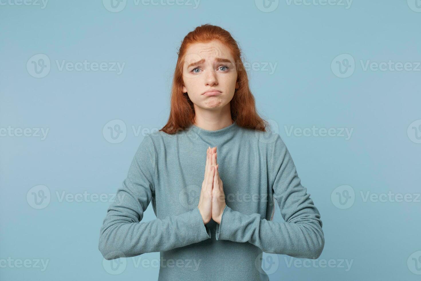 Red-haired girl prays asking for something. A teenager with a pitifully sad expression on her face folded her arms in front of herself showing a begging pleading gesture isolated over blue background photo